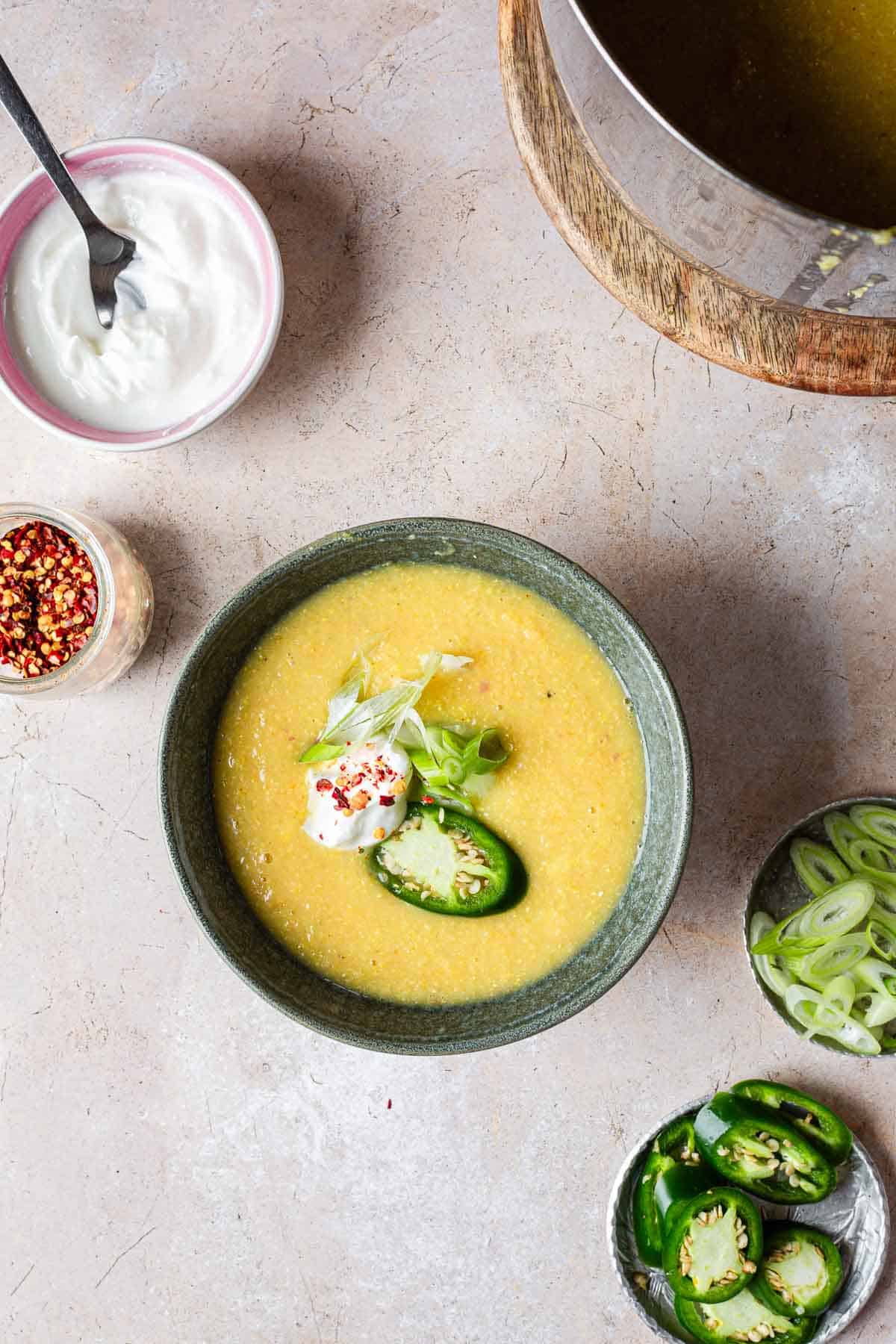 A bowl of creamy yellow corn soup garnished with sour cream, green onions, and slices of jalapeño, accompanied by bowls of spices and toppings on a light stone surface.