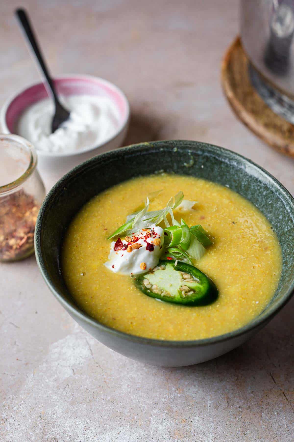 A bowl of smooth, yellow lentil and fresh corn soup garnished with a dollop of yogurt, a slice of green jalapeno, and fresh herbs. A spoon rests in a small bowl of yogurt nearby, and some spices are visible in the background.