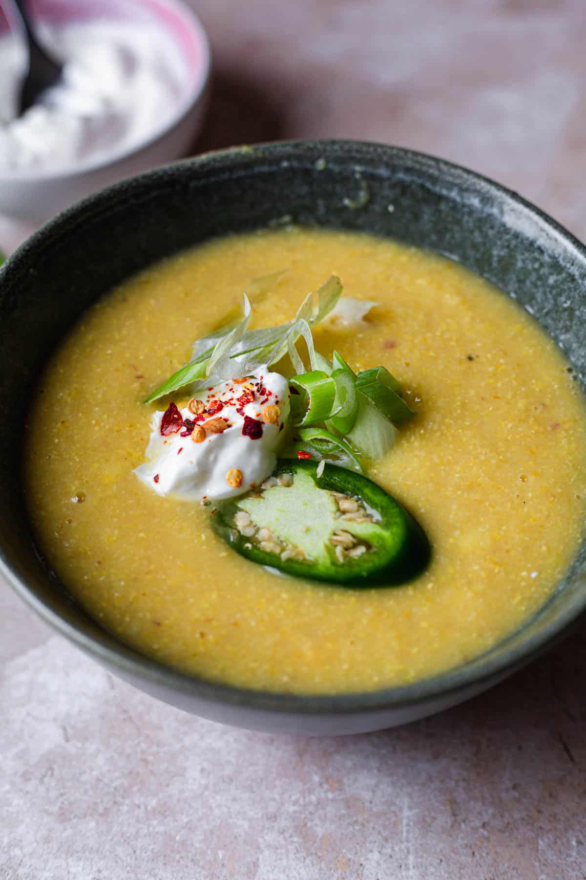 A bowl of smooth, creamy corn soup garnished with a dollop of sour cream, slices of green jalapeño, and a sprinkle of red chili flakes, served in a gray ceramic bowl.