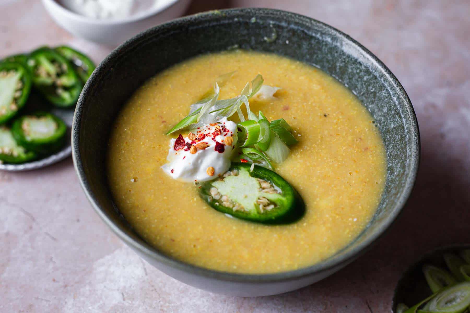 A bowl of creamy corn soup topped with a dollop of yogurt, slices of jalapeno, chopped herbs, and a sprinkle of red spices, served on a marble tabletop with additional jalapenos and a bowl of yogurt on the side.