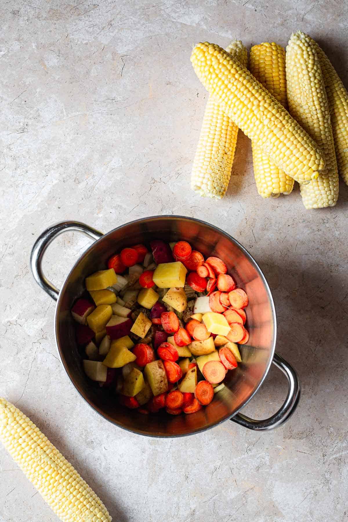 A stainless steel pot containing chopped carrots, potatoes, and spices on a textured gray surface, with whole corn cobs arranged on the side for an Easy Corn Soup.