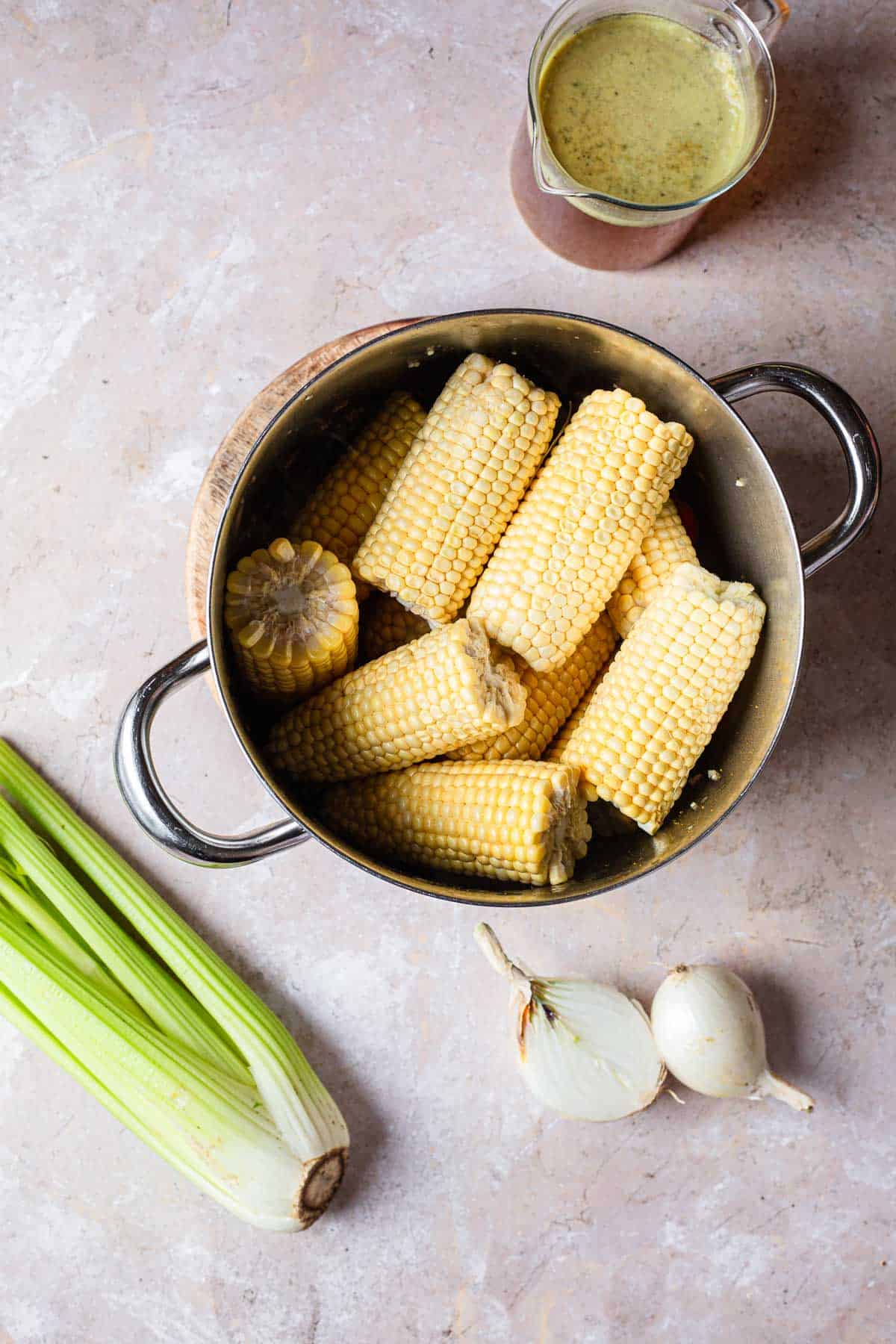 A pot filled with boiled fresh corn on the cob, accompanied by a gravy boat of sauce, onions, and leek on a textured surface.