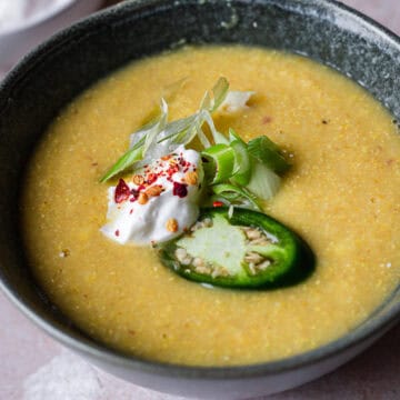 A bowl of creamy, blended fresh corn soup garnished with a dollop of cream, sliced green jalapeños, basil leaves, and a sprinkle of red pepper flakes. The soup has a smooth texture with visible specks of spices.