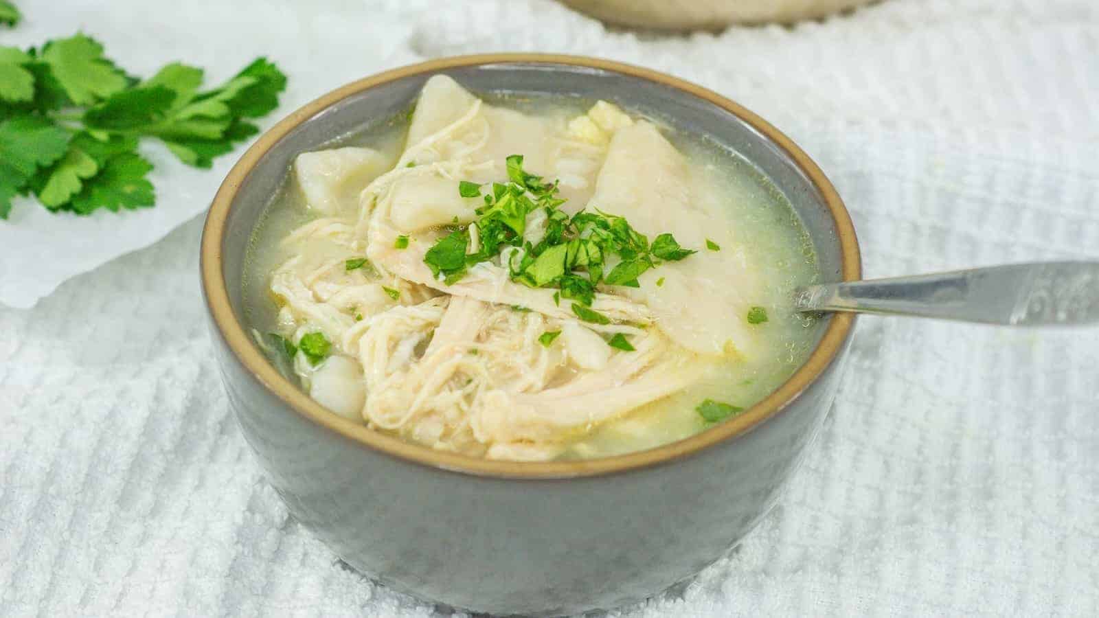 A bowl of irresistible chicken soup with dumplings, garnished with chopped parsley, on a white textured cloth, with a spoon on the side.