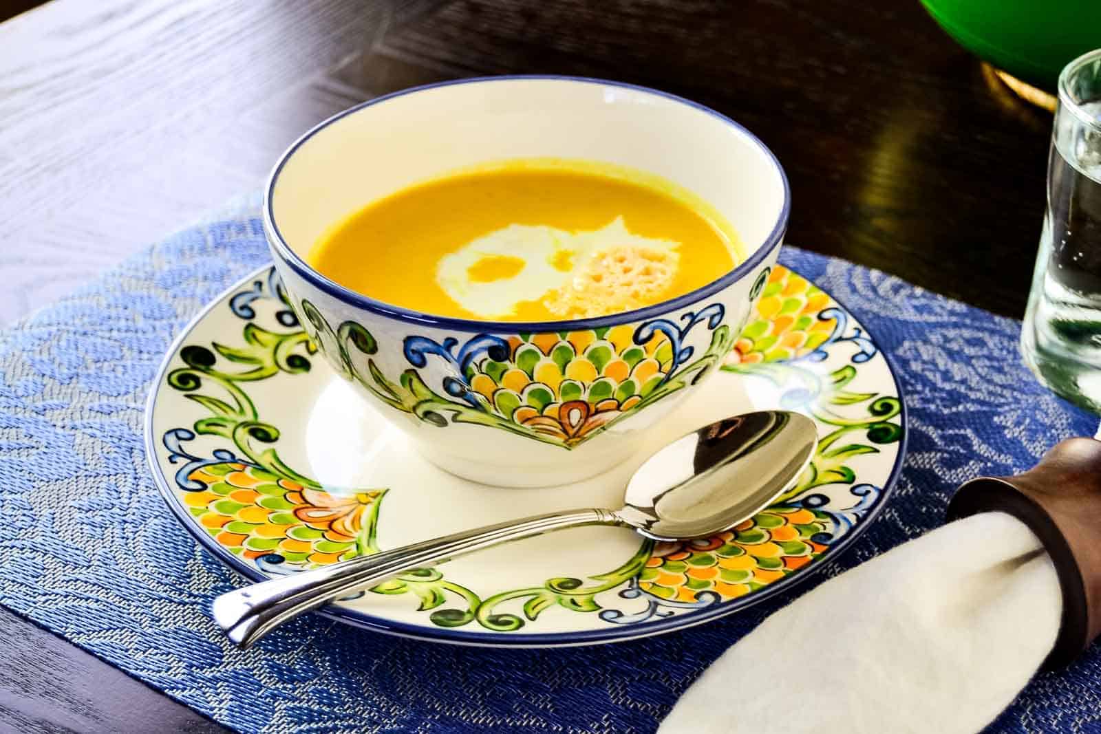A colorful ceramic bowl of creamy pumpkin soup, inspired by recipes from the '60s, on a wooden table, with a spoon to the side. The bowl is placed on a matching saucer and