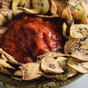 A ceramic plate filled with red sauce from old school recipes surrounded by a ring of baked plantain chips, presented on a light-colored surface.