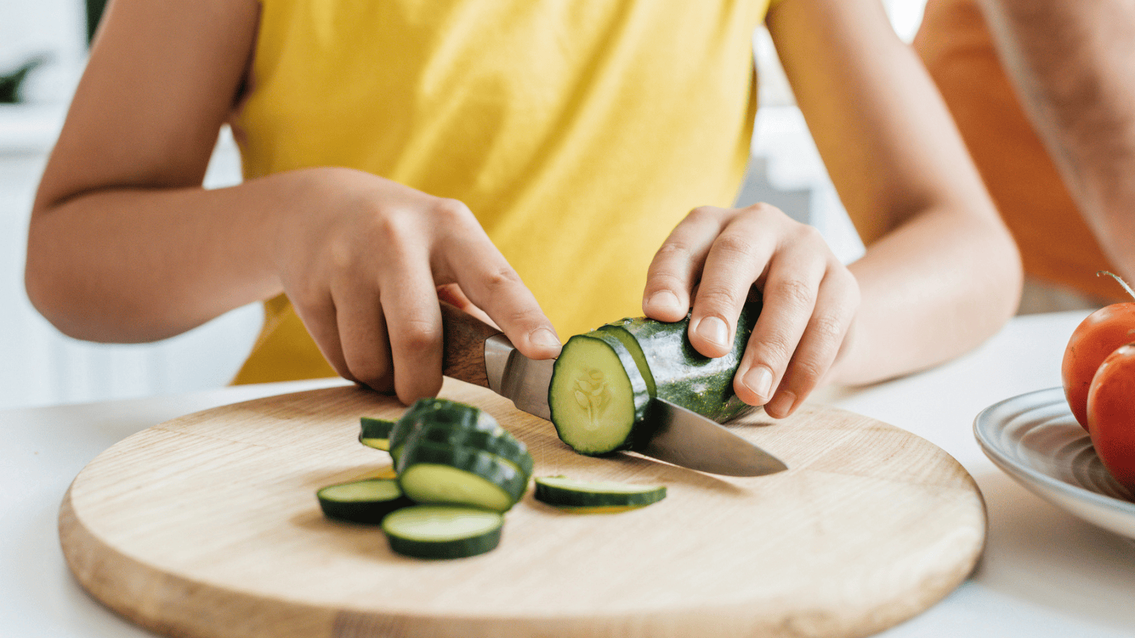 A person wearing a yellow shirt is slicing a cucumber on a wooden cutting board. The person's hands hold the cucumber and a knife. Some sliced cucumbers are on the board. There is a plate with tomatoes in the background.