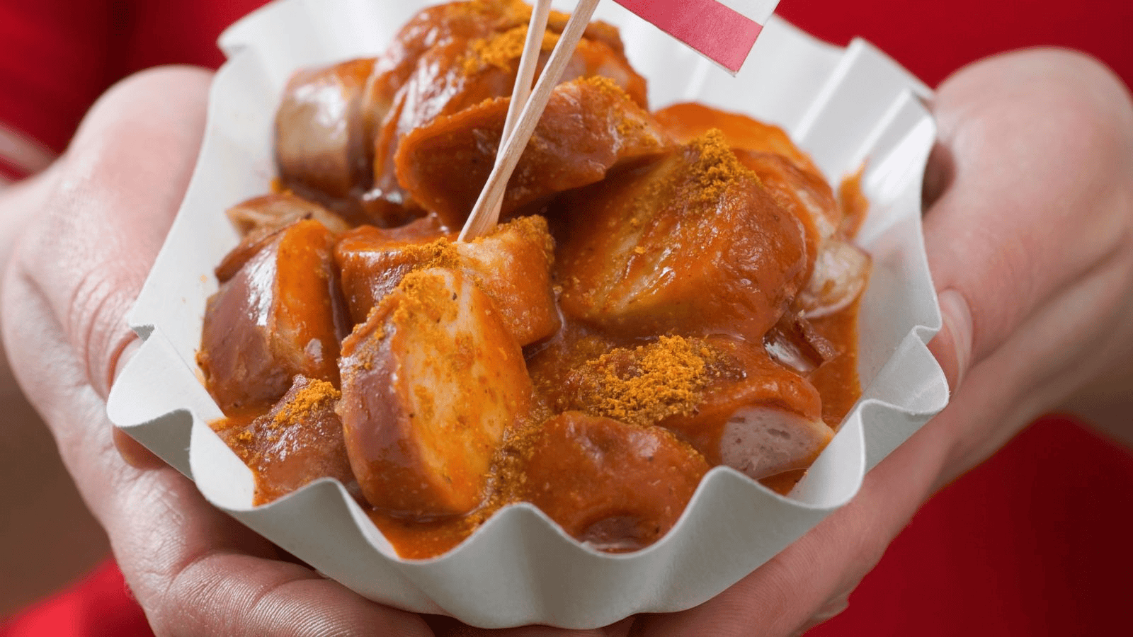 A person holding a tray of currywurst. The dish consists of sliced sausage covered in spiced ketchup and sprinkled with curry powder. The tray has a crimped edge, and a small paper flag and a toothpick are stuck into the sausage pieces.