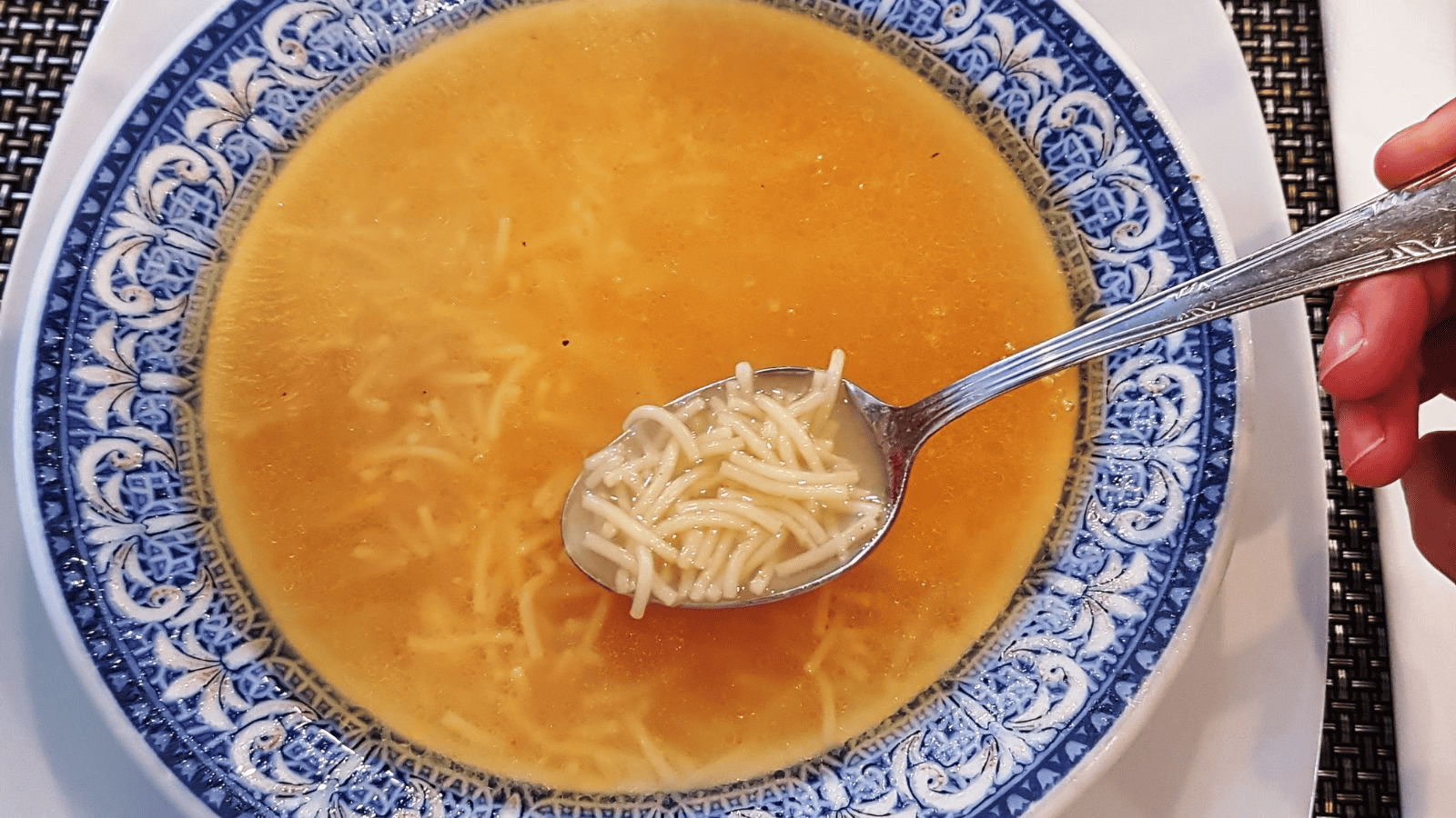 A bowl of noodle soup is placed on a white plate. The blue and white patterned bowl contains clear broth with thin noodles. A hand holds a spoon with noodles over the bowl. The background features a woven placemat.