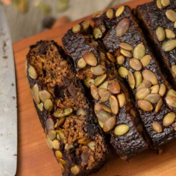 A loaf of dark-colored breakfast bread, topped with pumpkin seeds, is partially sliced on a wooden cutting board. A bread knife with remnants of crumbs is placed next to the loaf.