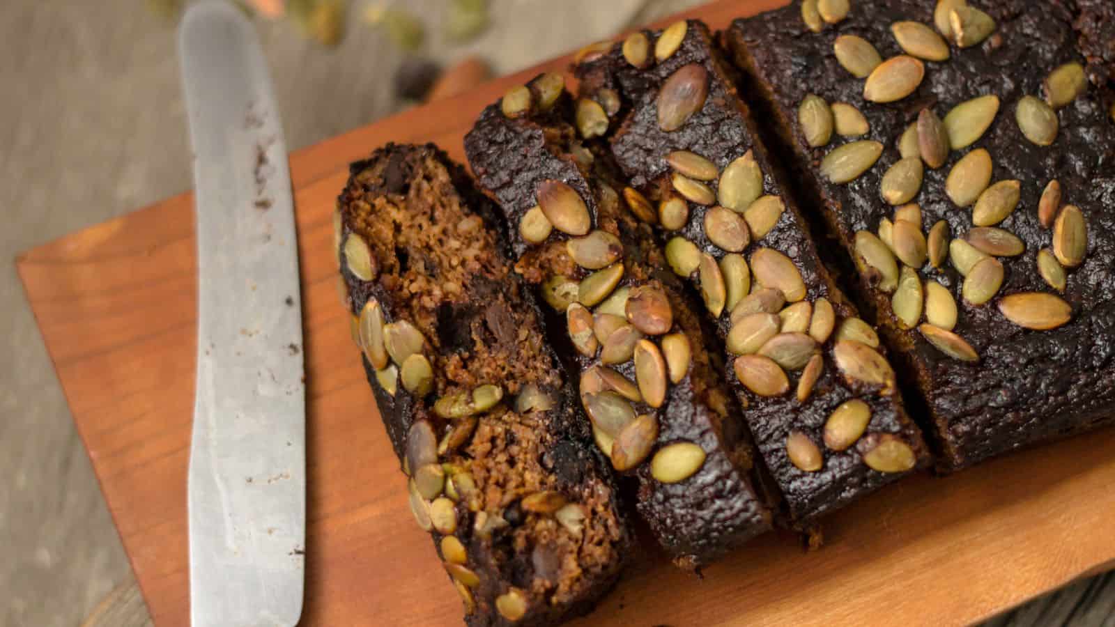 A loaf of dark-colored breakfast bread, topped with pumpkin seeds, is partially sliced on a wooden cutting board. A bread knife with remnants of crumbs is placed next to the loaf.