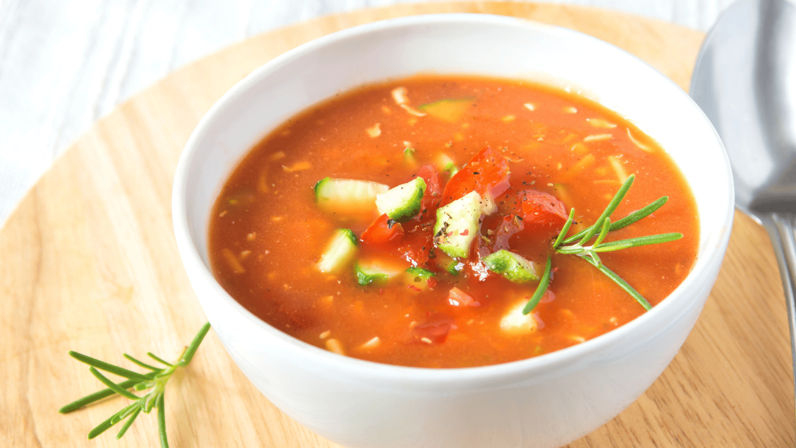 A white bowl filled with tomato-based gazpacho soup garnished with diced vegetables and a sprig of rosemary sits on a wooden surface. A spoon is placed to the right of the bowl.