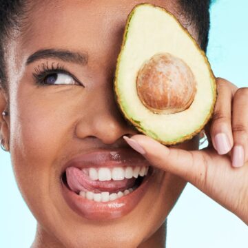 A joyful woman holding a halved avocado over her right eye, smiling broadly, against a light blue background.