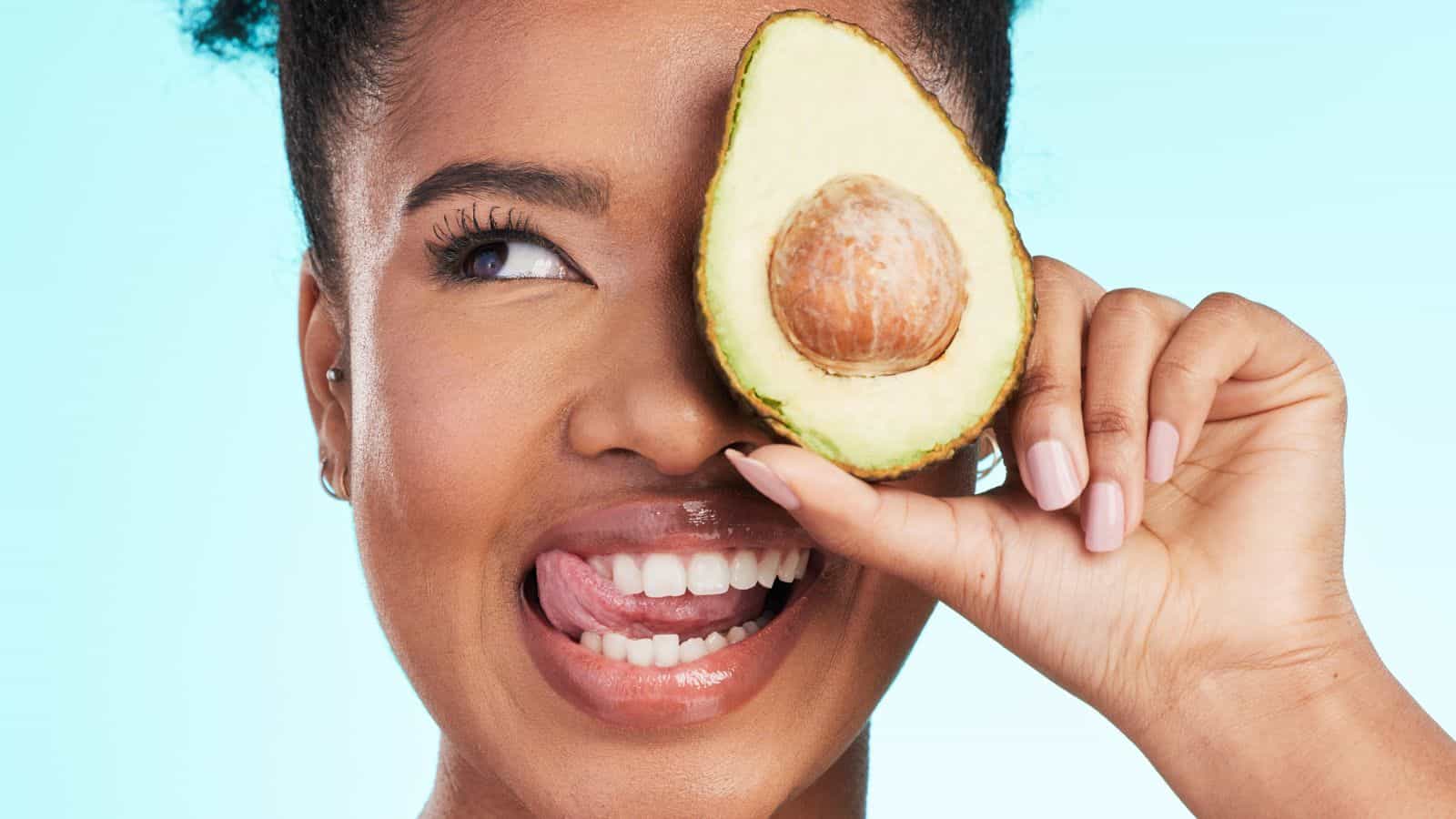 A joyful woman holding a halved avocado over her right eye, smiling broadly, against a light blue background.