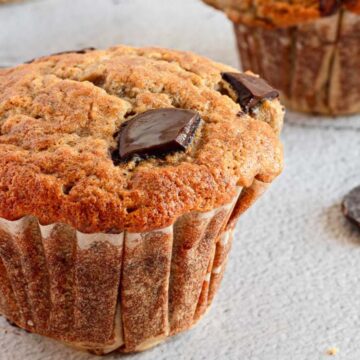 A close-up of a chocolate chip muffin on a light, textured surface, with another breakfast bread and scattered chocolate pieces in the background.