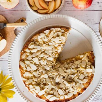 A top-down view of a table with a partially sliced apple-almond cake on a white plate. Sliced apples are on a cutting board and in a bowl. A fork and a plate with a piece of cake are beside the main plate. Yellow flowers and whole apples are also visible.