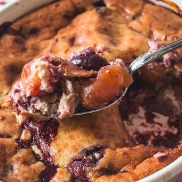 A close-up of a spoon scooping a portion of fruit cobbler from a large white baking dish. The cobbler has a golden-brown crust with visible chunks of fruit, and some of the filling is oozing through, resembling those beloved Retro Recipes. A portion is already missing from the dish.