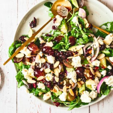 A plate of goat cheese salad with leafy greens, crumbled cheese, sliced almonds, red onions, and a variety of berries. The plate is garnished with two wooden spoons. A small bowl of red dressing is placed next to the plate, all set on a white wooden surface.