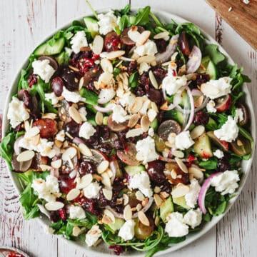 A plate of salad featuring mixed greens, sliced cucumbers, red onion, grapes, crumbled goat cheese, sliced almonds, and a light dressing. The goat cheese salad is served on a white wooden surface.