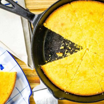 Golden cornbread in a cast iron skillet with one slice served on a white plate with blue detailing, set on an auto draft wooden table.