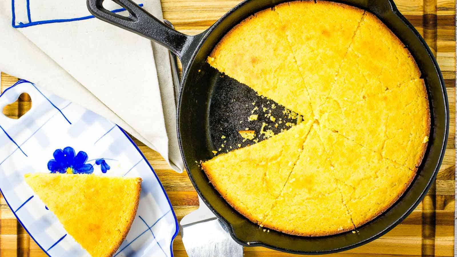 Golden cornbread in a cast iron skillet with one slice served on a white plate with blue detailing, set on an auto draft wooden table.