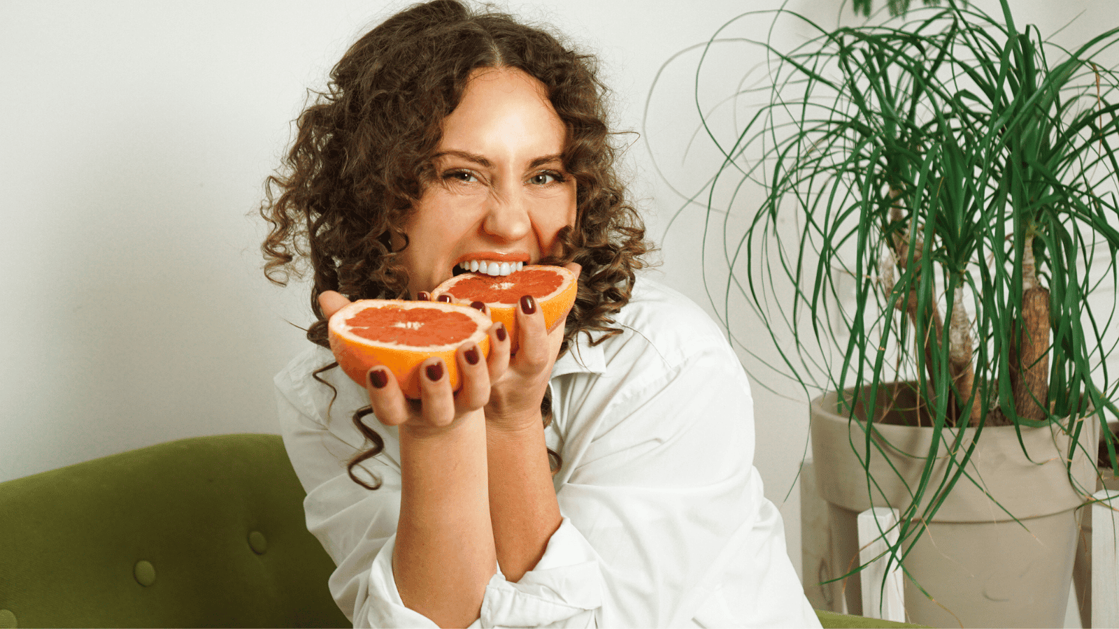 A person with curly hair and a white shirt is smiling and holding two halves of a grapefruit. They are sitting on a green couch. There is a tall potted plant with long, thin leaves in the background.