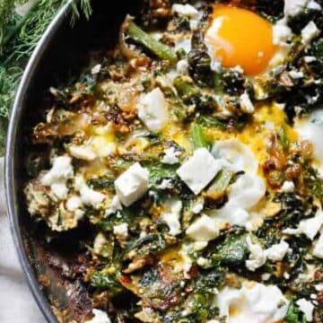 A skillet contains a cooked dish with two sunny-side-up eggs atop a mixture of spinach, onions, and crumbled feta cheese. Fresh herbs, likely dill, are visible on the side, and a small bowl contains additional cubed feta cheese.