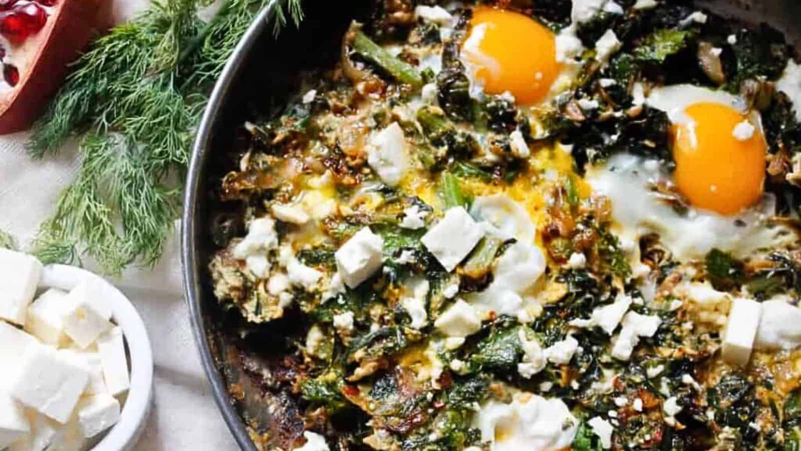 A skillet contains a cooked dish with two sunny-side-up eggs atop a mixture of spinach, onions, and crumbled feta cheese. Fresh herbs, likely dill, are visible on the side, and a small bowl contains additional cubed feta cheese.