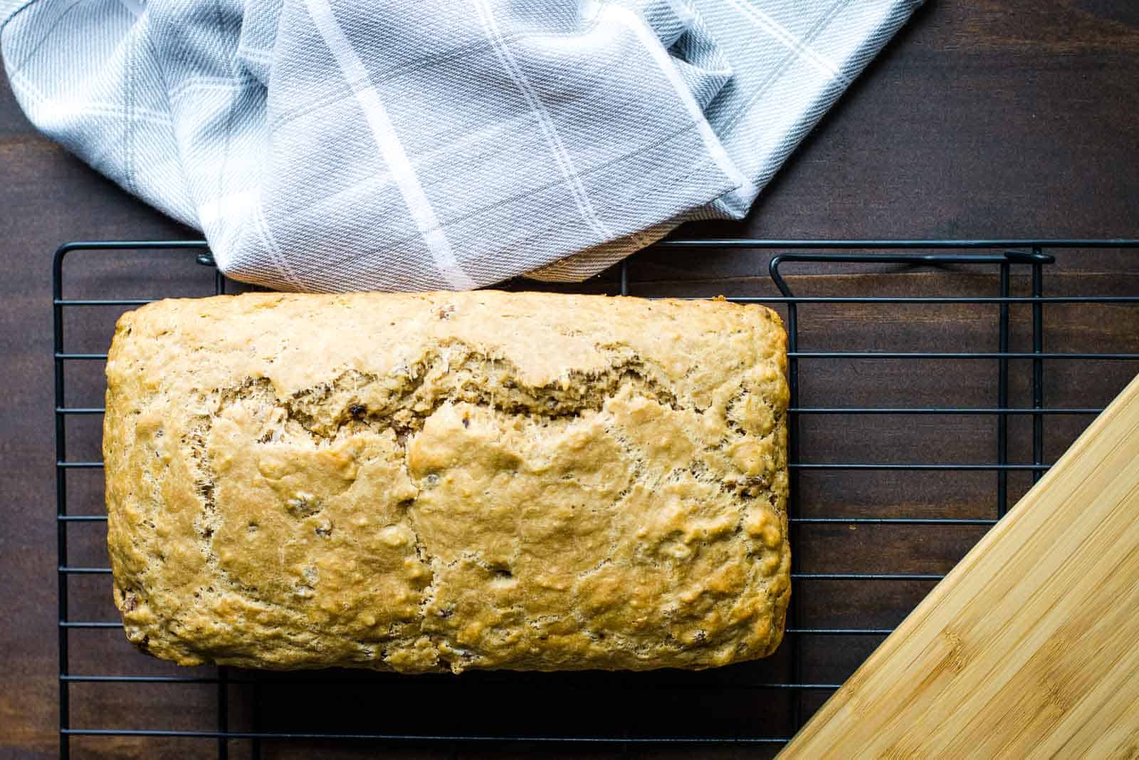 A loaf of bread cooling on a black wire rack. There is a white and gray checkered cloth partially covering the top corner of the rack, and a light-colored wooden cutting board is positioned on the right side of the image, perfect for slicing into delicious breakfast breads.