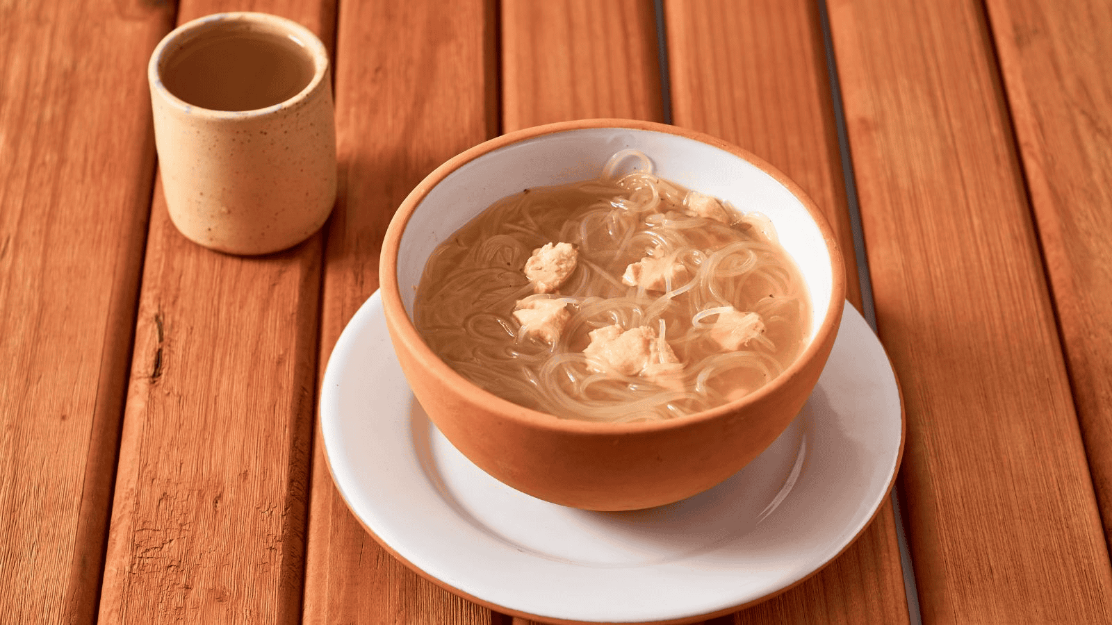 A bowl of noodle soup with pieces of meat is placed on a white saucer atop a wooden table. A beige cup filled with a light brown liquid is located to the left of the bowl.