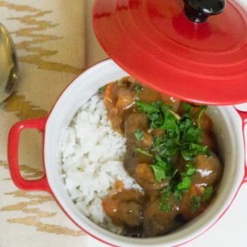 A red-lidded pot containing a meal of white rice, stew with chunks of meat, and garnished with chopped parsley, reminiscent of Grandpa's recipes. A silver spoon is placed on a beige patterned cloth beside the pot, begging for more servings.