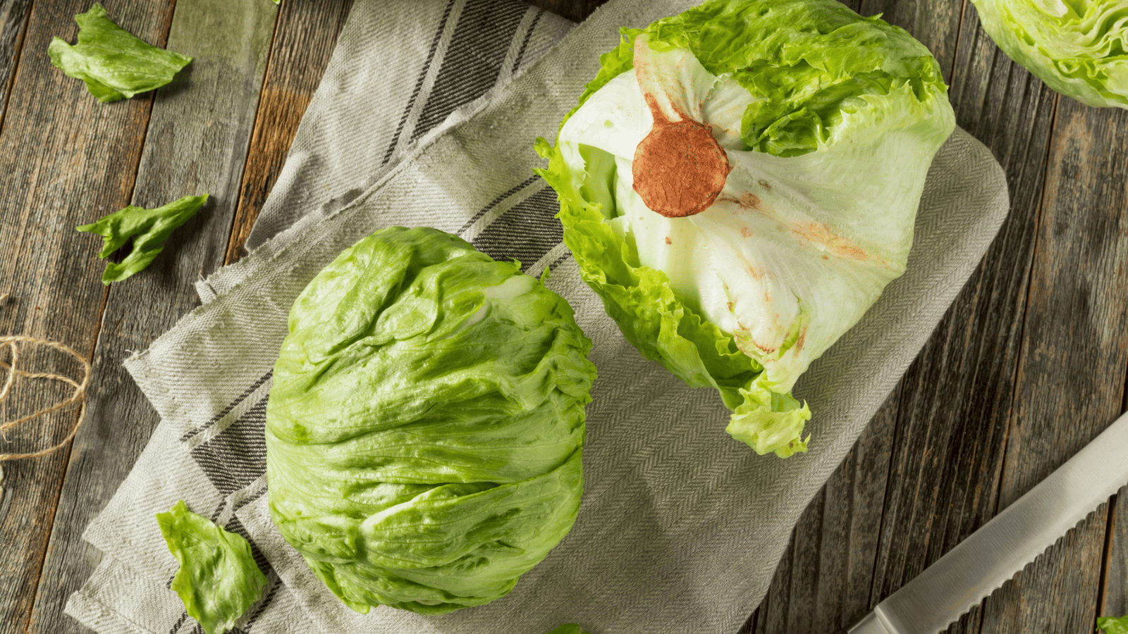 Two heads of iceberg lettuce are placed on a gray and white cloth on a rustic wooden surface. One lettuce head is whole, while the other is partially cut, revealing the core. A knife rests nearby along with a few scattered lettuce leaves.