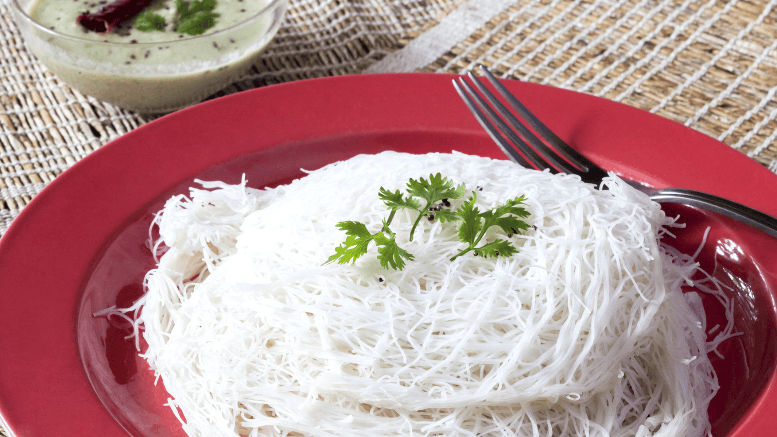 A plate with a serving of white string hoppers garnished with cilantro, accompanied by a fork. A bowl of chutney with a garnish and a red chili is also partially visible in the background on a woven placemat.