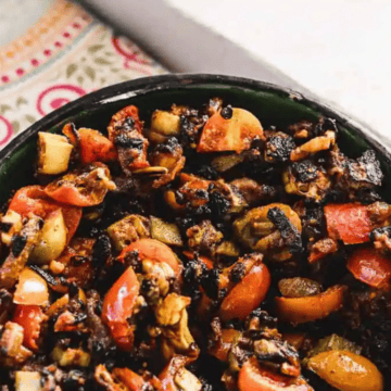 A bowl filled with a mixture of charred vegetables and small pieces of seafood sits on a colorful, patterned tablecloth. The dish includes chopped tomatoes, onions, and possibly clams or mussels. There is a white windowsill in the background.