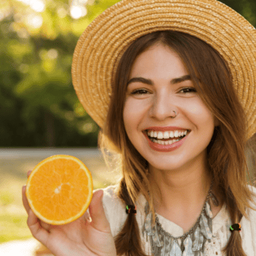 A smiling young woman wearing a straw hat holds up half an orange. She is outdoors in a park-like setting with greenery in the background. She has two braids and wears a light-colored top.