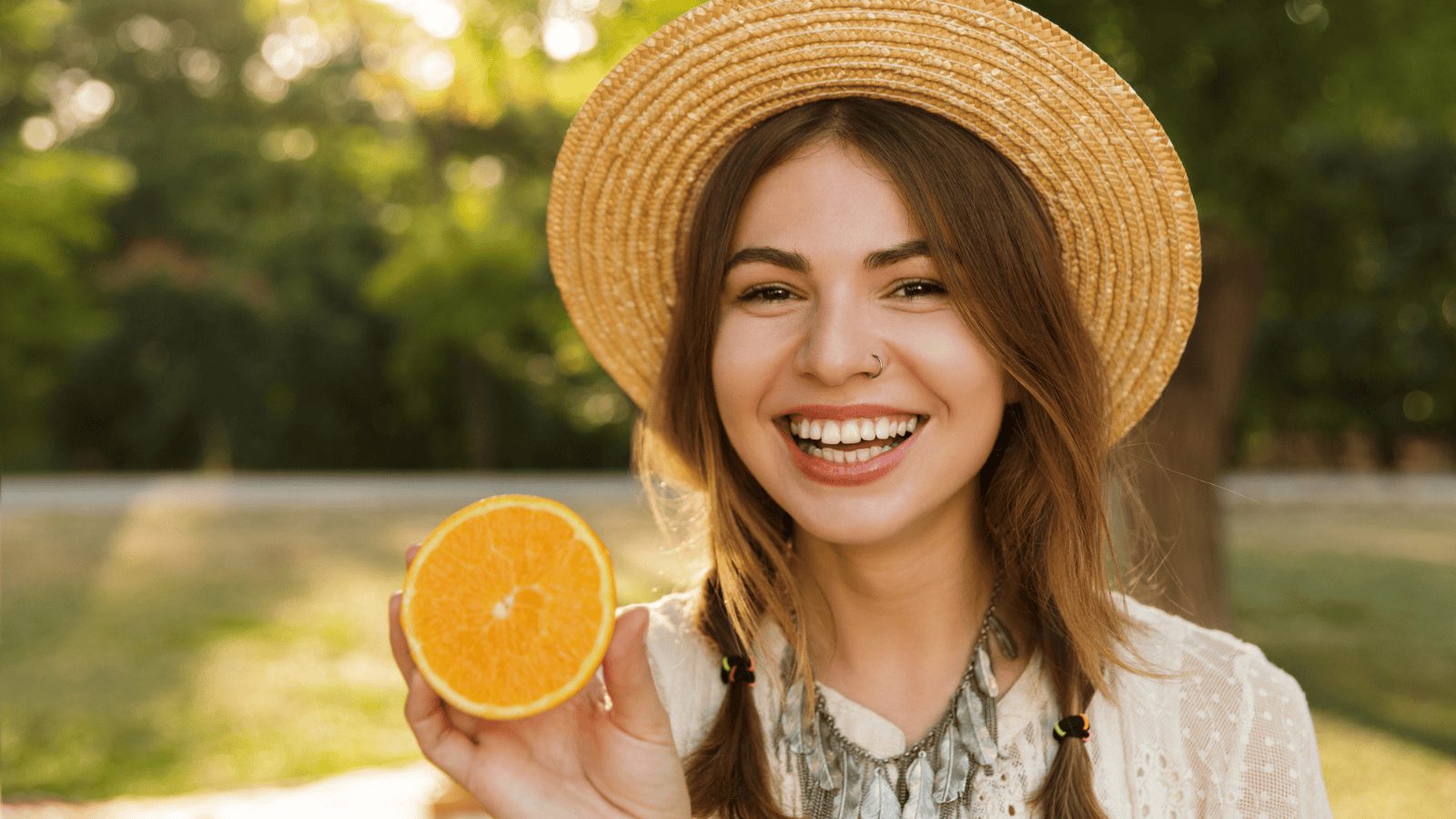 A smiling young woman wearing a straw hat holds up half an orange. She is outdoors in a park-like setting with greenery in the background. She has two braids and wears a light-colored top.
