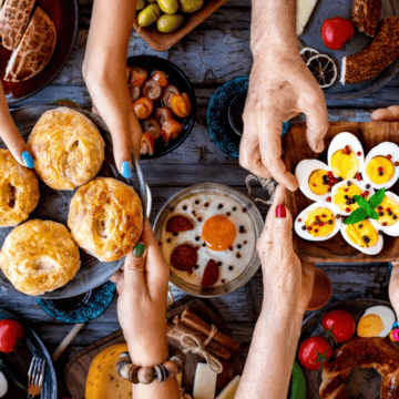 A variety of foods are displayed on a table. Four hands hold a plate of baked pastries, while two hands hold a tray of halved boiled eggs with toppings. The table includes dishes like fried eggs, tomatoes, pickles, and drinks in blue cups.