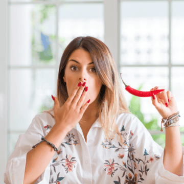 A woman with long hair stands indoors, holding a red chili pepper in one hand and covering her mouth with the other. She wears a white floral blouse and has a surprised expression. There is a bright background with large windows.