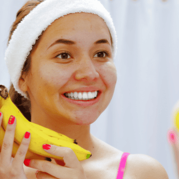 A woman with a white headband and red-painted nails is standing in front of a mirror, smiling, and holding bananas in both hands. She appears to be in a bathroom with a white curtain in the background.