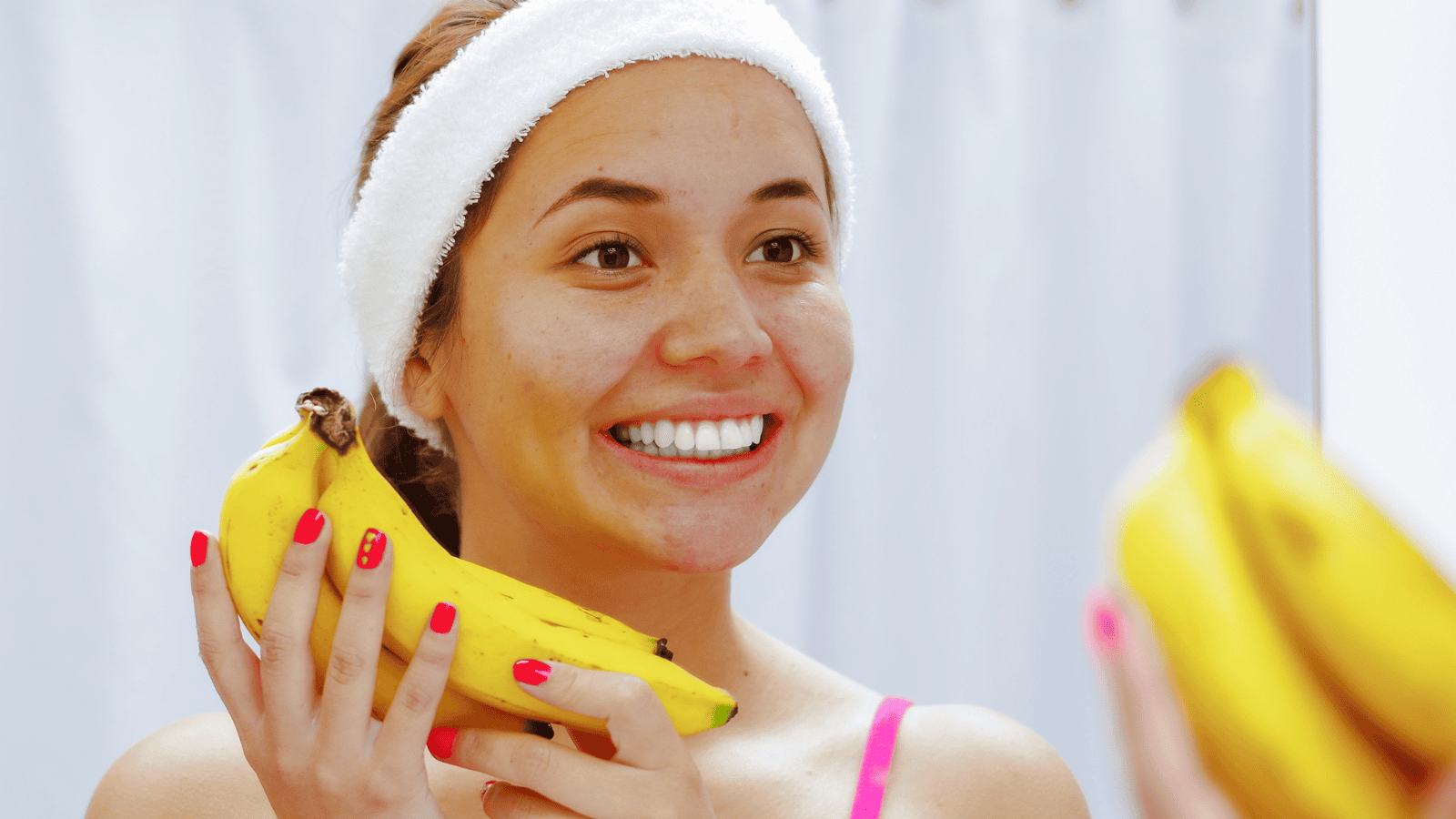 A woman with a white headband and red-painted nails is standing in front of a mirror, smiling, and holding bananas in both hands. She appears to be in a bathroom with a white curtain in the background.