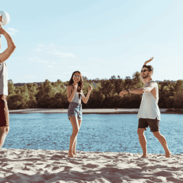 A group of five young adults is playing beach volleyball by a lake. One person is about to serve the ball while the others are prepared to play. Trees and clear skies are in the background. Everyone is casually dressed and appears to be enjoying the outdoor activity.