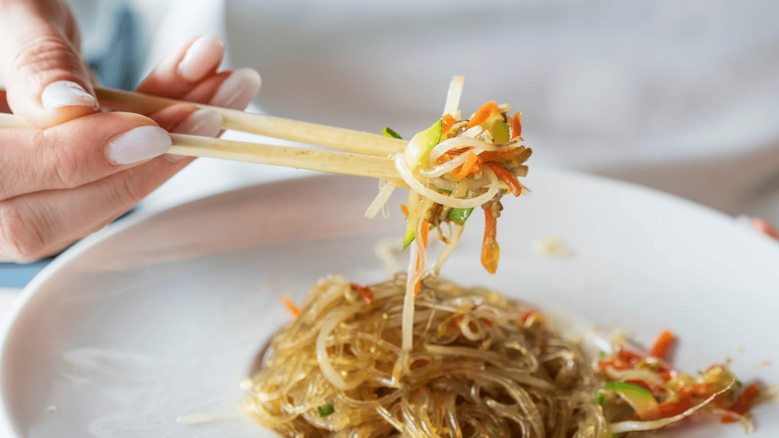 A person holds chopsticks with a portion of vegetable noodles above a white plate containing more noodles and vegetables. The noodles appear translucent and are mixed with shredded carrots, zucchini, and bean sprouts.