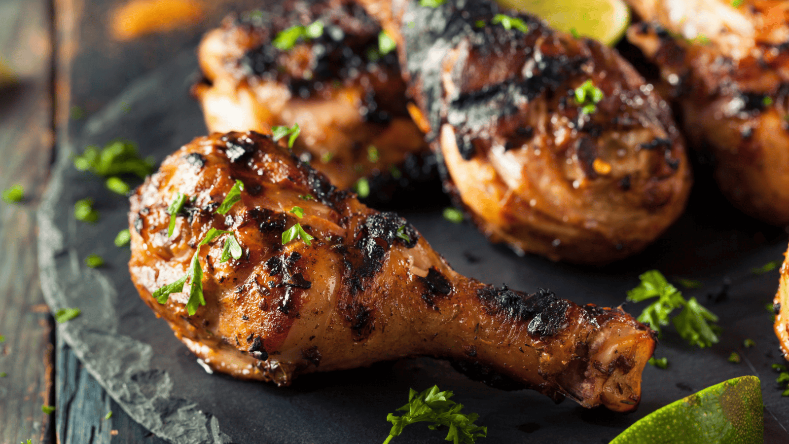 Grilled chicken drumsticks are placed on a dark surface. The drumsticks are charred and garnished with chopped green herbs. Lime wedges are partially visible in the background. The surface is sprinkled with herbs and spices.