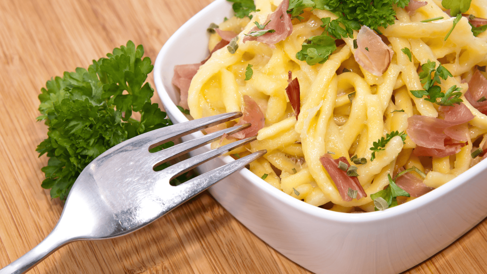 A square white bowl filled with pasta, garnished with pieces of prosciutto and fresh parsley. A metal fork rests on the edge of the bowl. The bowl is placed on a wooden surface.