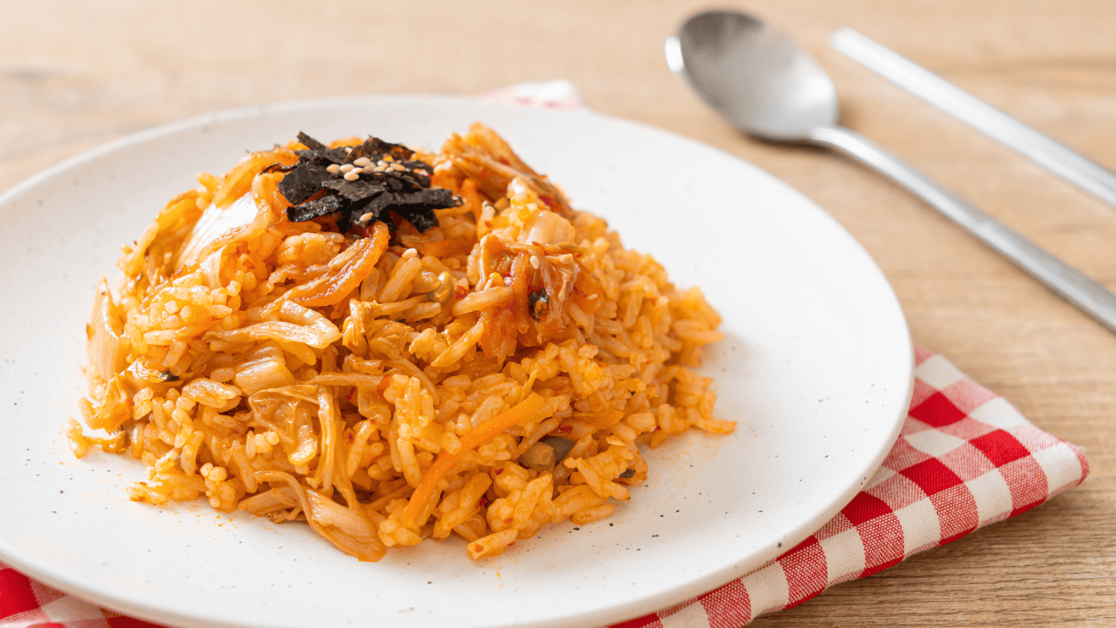 A plate of kimchi fried rice garnished with seaweed and sesame seeds on a white plate. The plate rests on a red and white checkered napkin, and a spoon and fork are placed beside it on a wooden surface.
