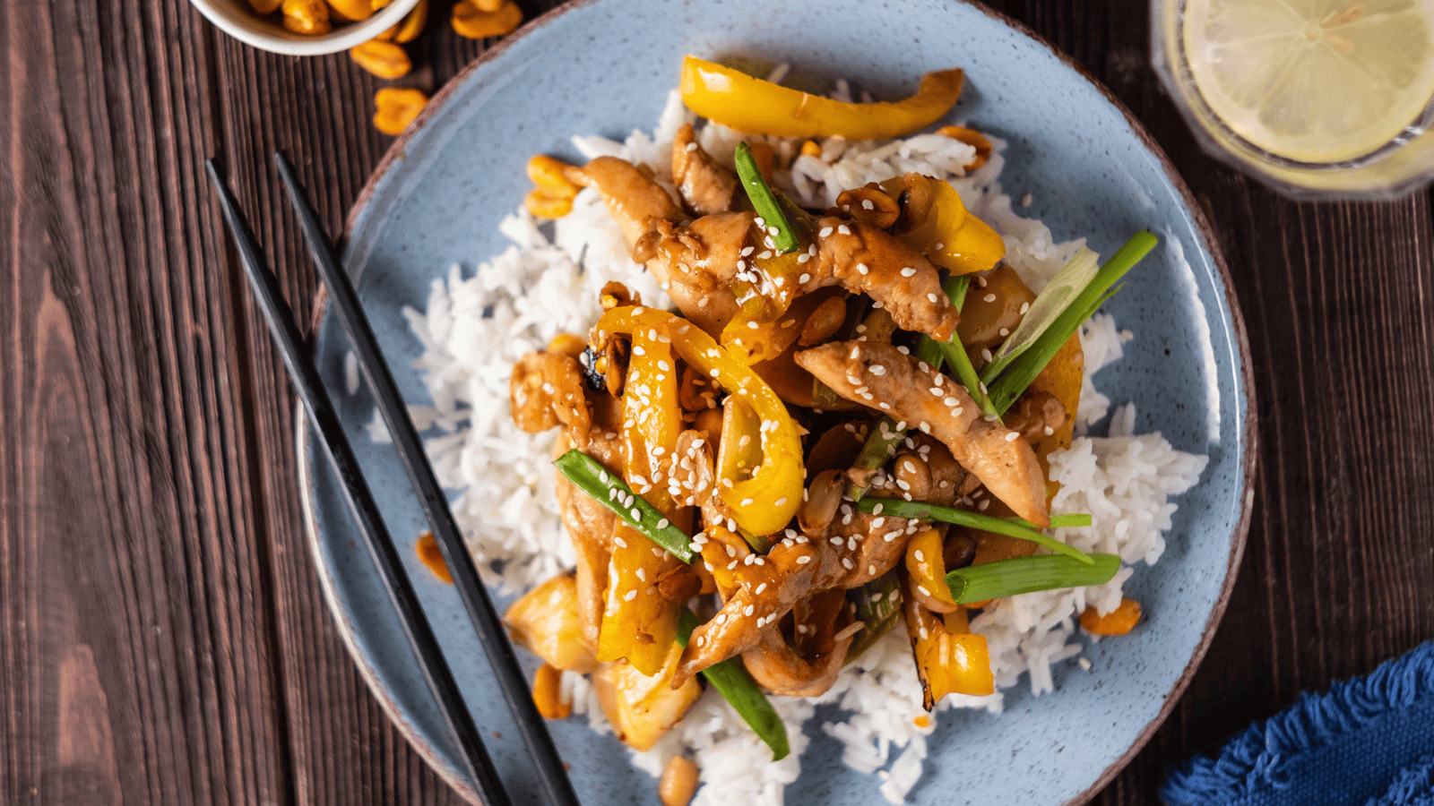 A plate of stir-fried chicken and vegetables, including bell peppers and green onions, served on a bed of white rice. Black chopsticks are placed across the plate. A lemon slice is visible in a glass of water at the upper right corner.