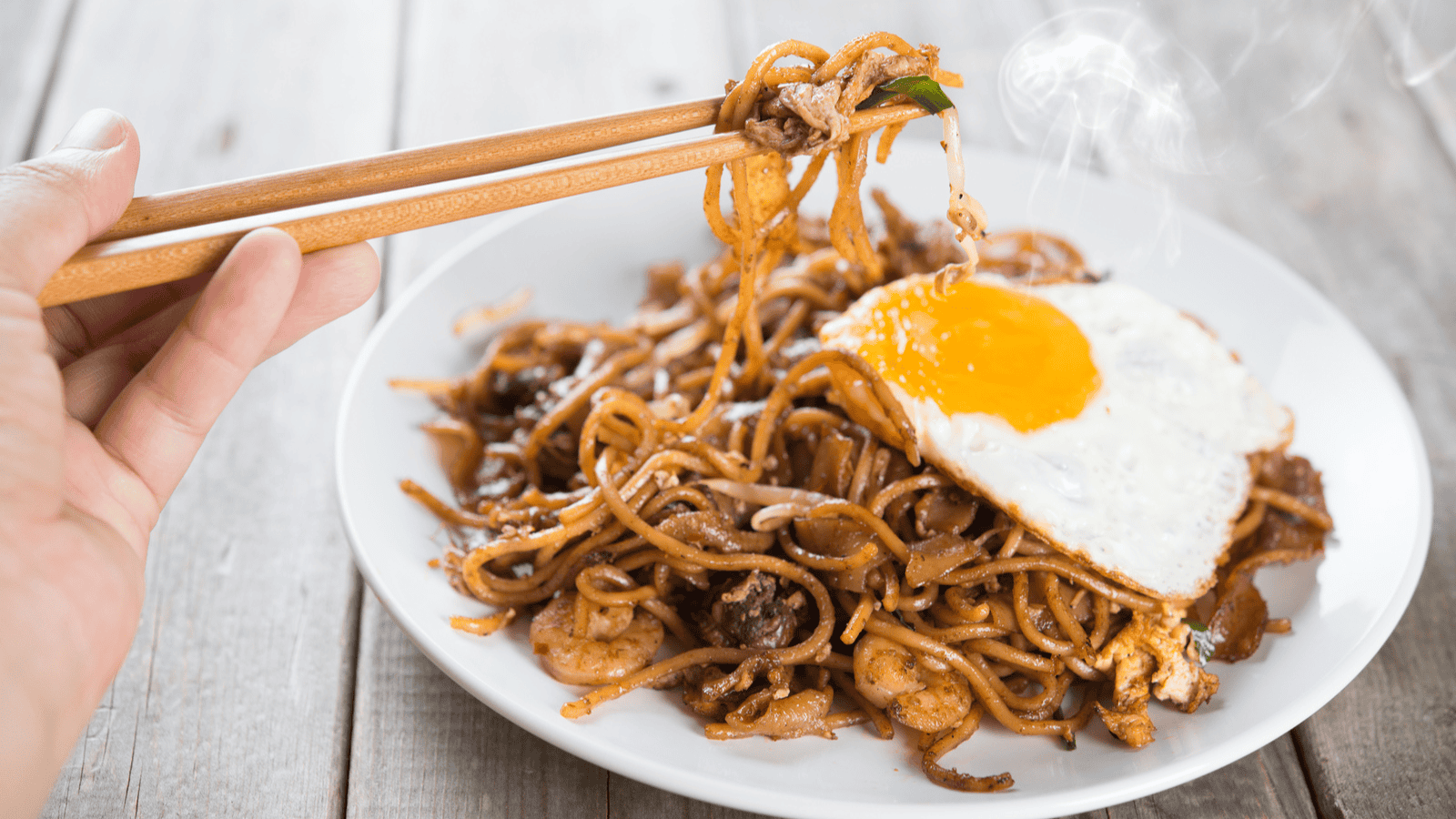 A hand holding chopsticks is lifting noodles from a plate of stir-fried noodles topped with a sunny-side-up egg. The dish contains shrimp, meat, and vegetables, and steam is rising from the hot food. The plate is on a wooden surface.