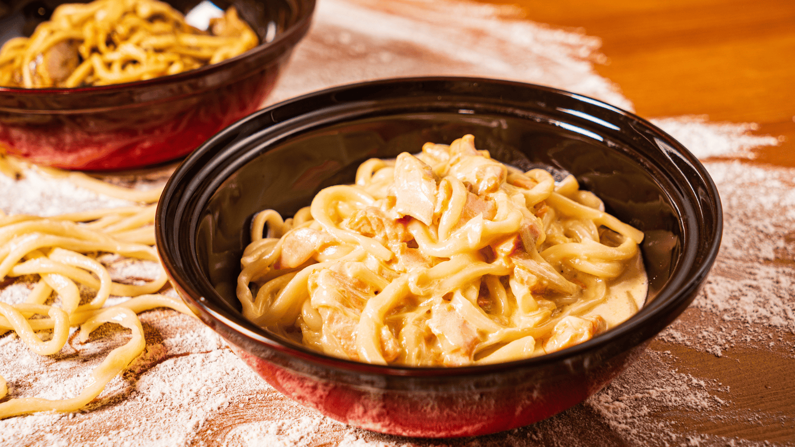 A close-up view of two bowls filled with creamy pasta topped with pieces of chicken. The bowls are placed on a wooden surface dusted with flour, and some pasta strands are scattered around. The dish appears to be freshly prepared.