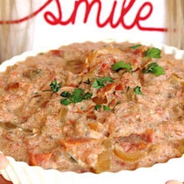 A person holding a white bowl of creamy tomato and onion dip from '60s recipes, garnished with parsley, with the word "smile" written in red in the background.