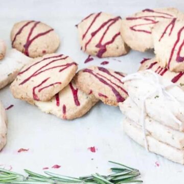 A group of cookies, reminiscent of grandma faves, is arranged on a white surface. Some cookies have red icing drizzled on top, while others are plain. A small bundle of plain cookies is tied with a white string, and a sprig of rosemary is placed nearby.